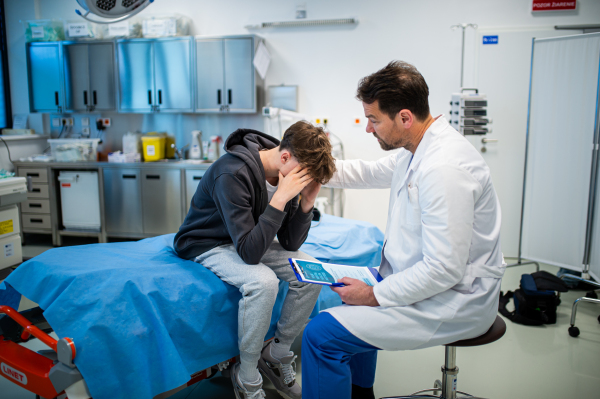 Depressed teenage boy patient sitting on hospital bed, talking with caring doctor.