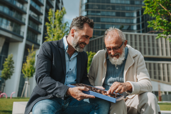 Senior dad and adult son sharing a box of sweets, chocolates on park bench. Fathers day.