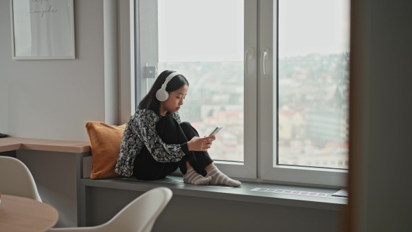 Young japanese girl with headphones scrolling her smartphone while listening to music next to window.