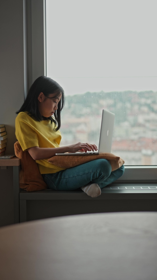 Young Japanese woman sitting by the window with a laptop, writing and working from home in natural light. Concept of remote work, creativity, freelance lifestyle, and online education.