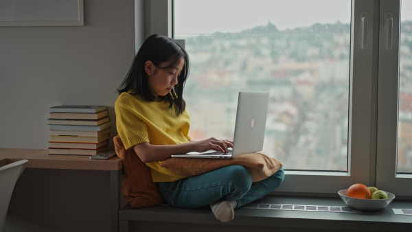 Young Japanese woman sitting by the window with a laptop, writing and working from home in natural light. Concept of remote work, creativity, freelance lifestyle, and online education.