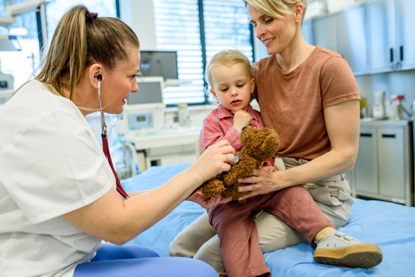 Pediatrician demonstrates stethoscope on teddy bear to calm toddler boy.