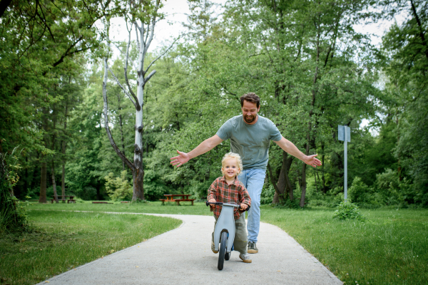 Toddler riding balance bike with dad cheering behind and running behind him.
