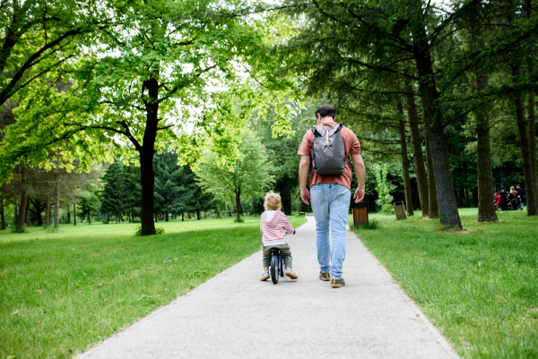 Rear view of father walking with his son balance bike across park