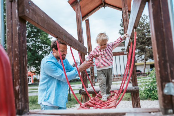 Toddler crossing playground obstacle with dad's support, weekend activity.