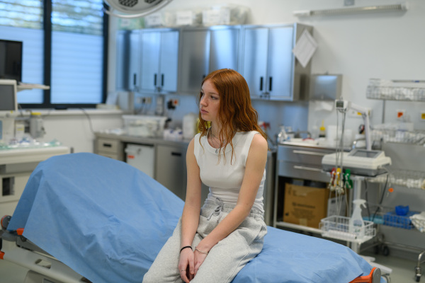 Teenage girl waiting for medical examination in hospital, sitting on examination table.