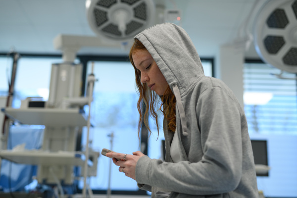 Teenage girl in hoodie waiting for medical examination in hospital, scrolling on phone.