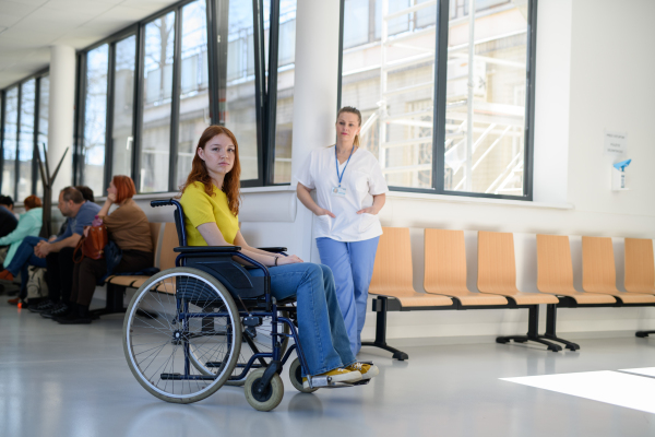 Nurse with young female patient in wheelchair at hospital, modern clinic.