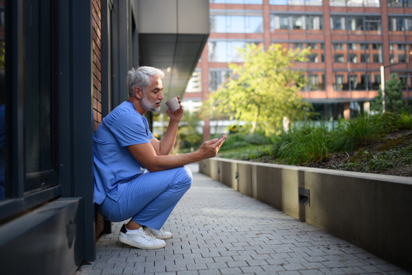 Healthcare worker in scrubs sitting outside hospital and checking phone during break.