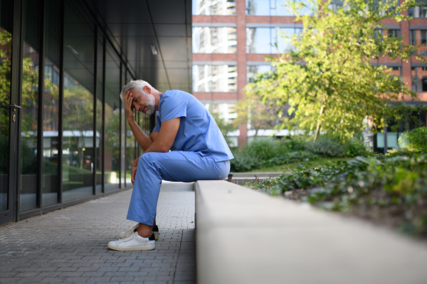 Frustrated and burnt-out doctor sitting outside the hospital, feeling down. Mental health problems.