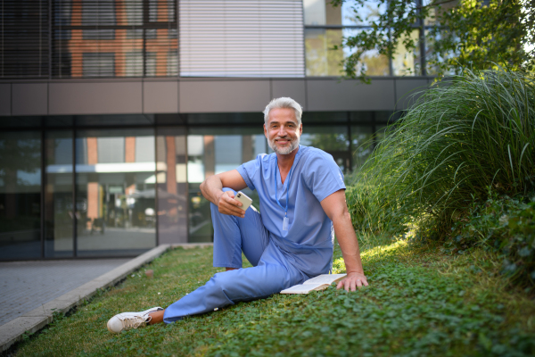 Healthcare worker resting in park outside hospital and holding smartphone.