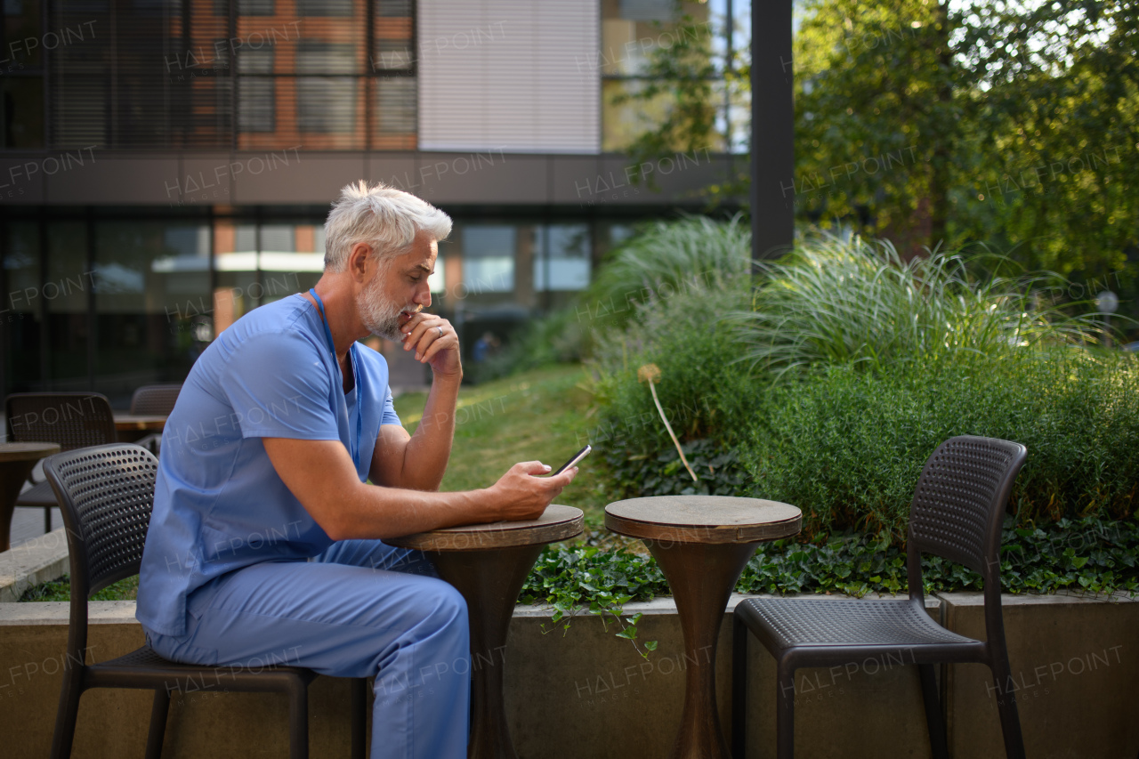Healthcare worker in scrubs sitting outside hospital and checking phone during break.