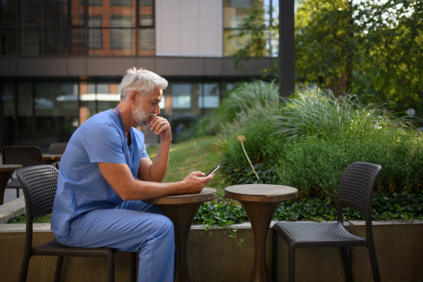 Healthcare worker in scrubs sitting outside hospital and checking phone during break.
