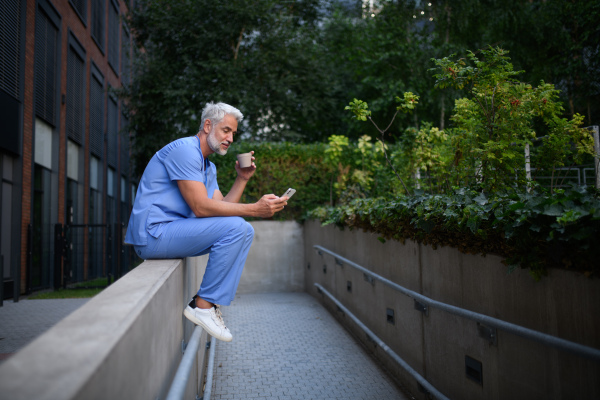 Healthcare worker in scrubs sitting outside hospital and checking phone during break.