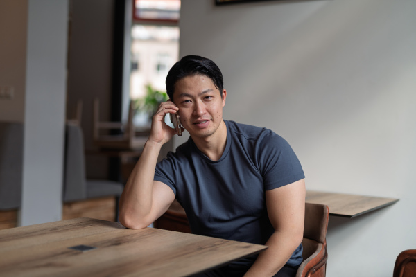 Young asian man talking on smartphone, sitting at empty table.