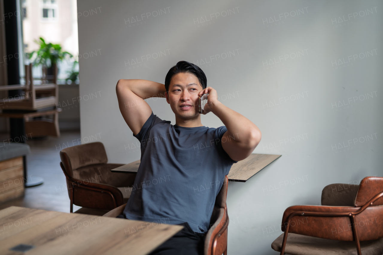 Confident young man having phone call at cafe table, sitting with hands behind head.