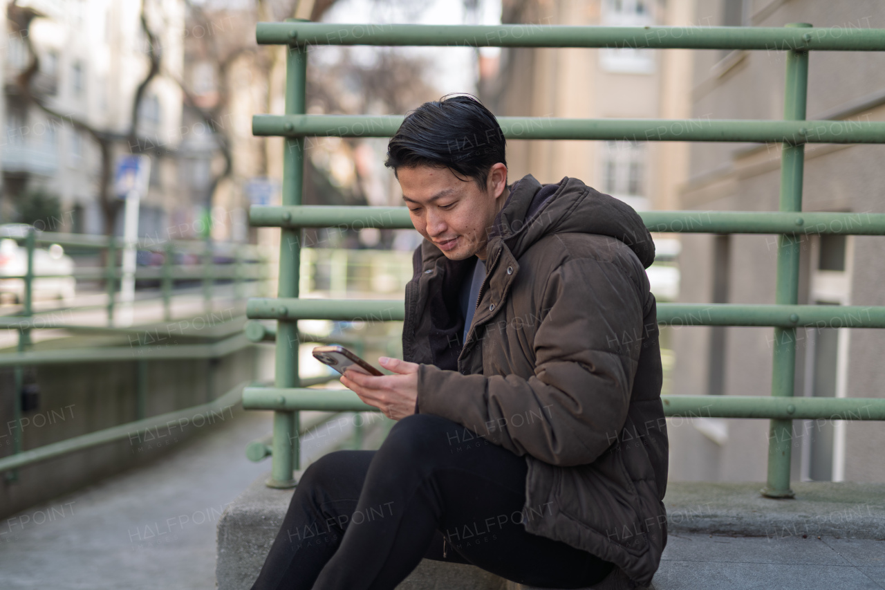 Young Asian man sitting alone on street ledge scrolling on smartphone.