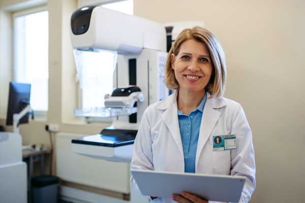 Smiling doctor standing next to mammogram machine in hospital. Preventive breast cancer screening.