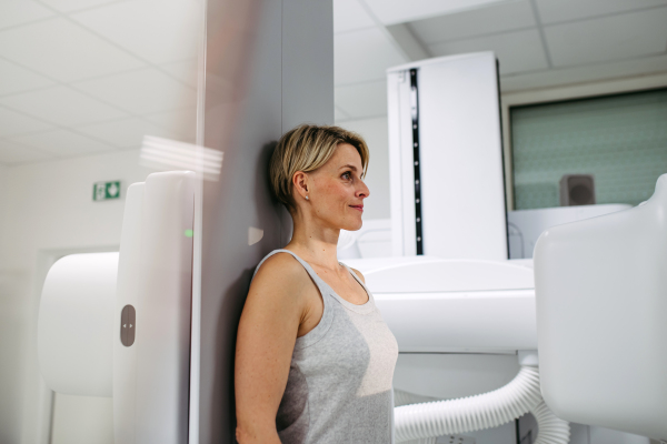 Female patient undergoing dynamic X-ray scan using tilting fluoroscopy wall unit, radiographic table with wall stand.
