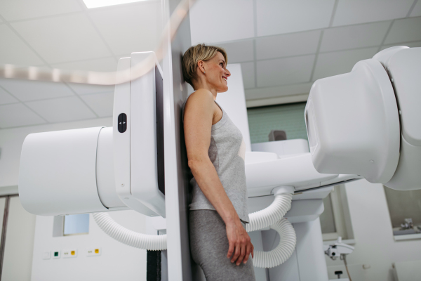 Female patient undergoing dynamic X-ray scan using tilting fluoroscopy wall unit, radiographic table with wall stand.