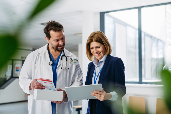 Female medical sales representative presenting medication to doctor, standing in hospital hallway.