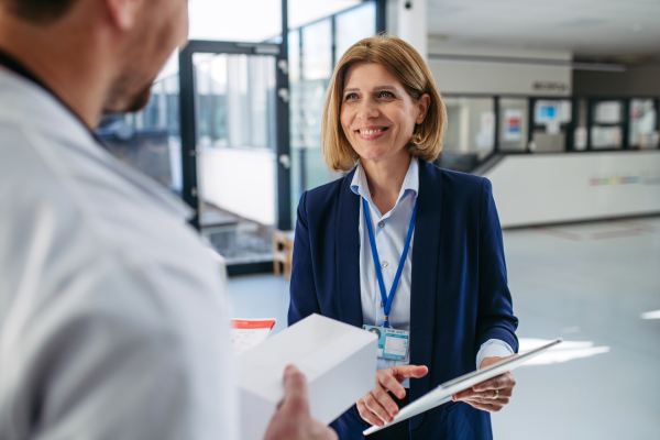 Female medical sales representative presenting medication to doctor, standing in hospital hallway.