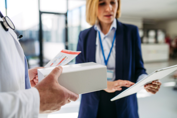 Female medical sales representative presenting medication to doctor, standing in hospital hallway.