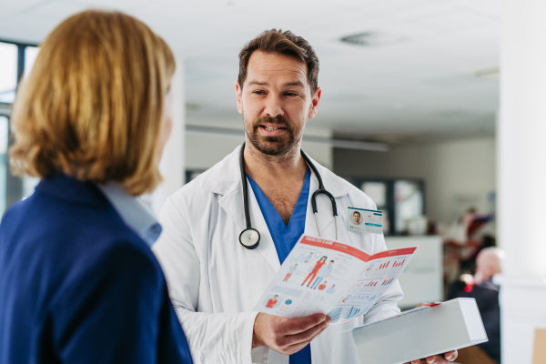 Female medical sales representative presenting medication to doctor, standing in hospital hallway.