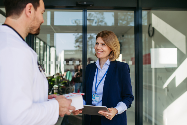 Female medical sales representative presenting medication to doctor, standing in hospital hallway.