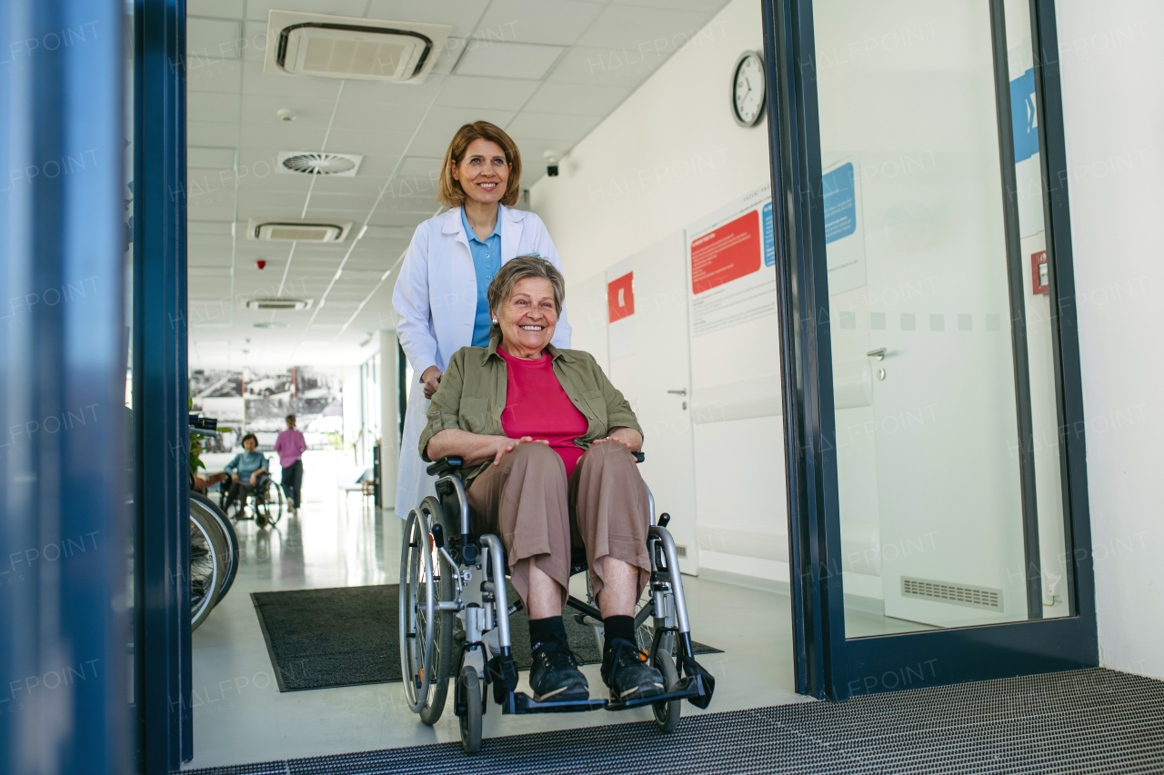 Friendly doctor pushing elderly patient in wheelchair across hospital hallway, Senior health care.