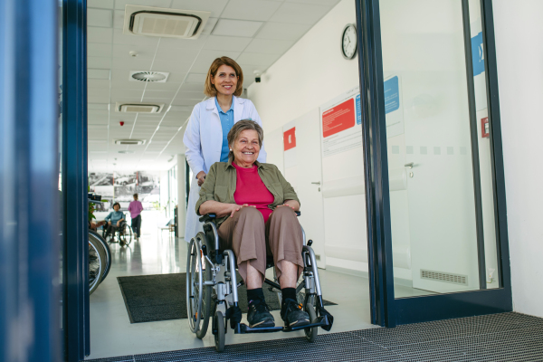 Friendly doctor pushing elderly patient in wheelchair across hospital hallway, Senior health care.