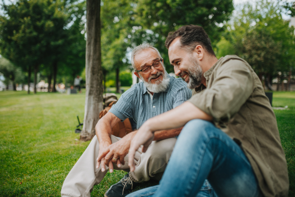 Father and adult son sitting on grass in park, sharing a story from the past. Fathers day concept.