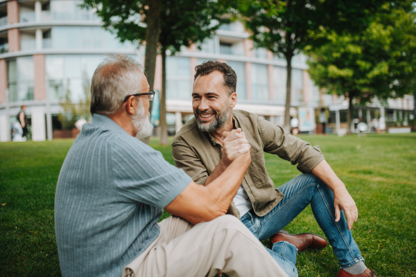 Father and adult son sitting on grass in park, sharing a story from the past. Fathers day concept.