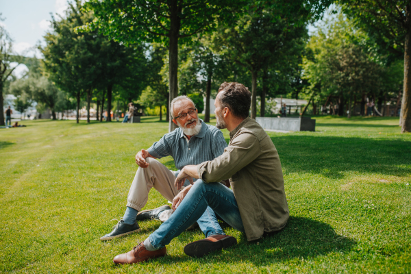 Father and adult son sitting on grass in park, sharing a story from the past. Fathers day concept.