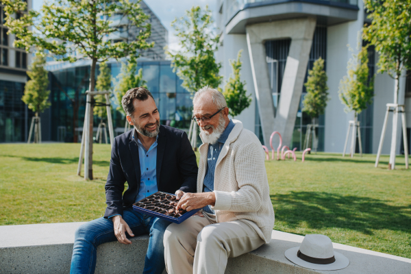Senior dad and adult son sharing a box of sweets, chocolates on park bench. Fathers day.