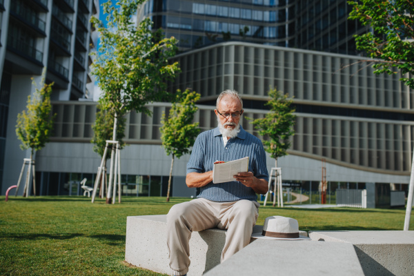 Senior man reading magazine or book in city park.