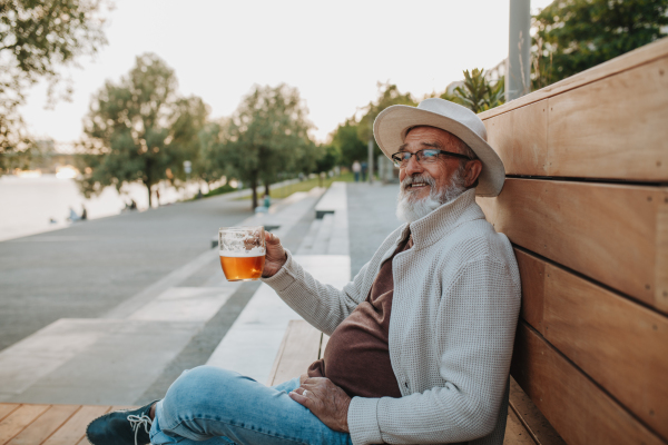 Senior man enjoying beer on sunny riverside promenade during sunset.