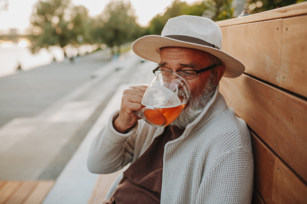 Senior man enjoying beer on sunny riverside promenade during sunset.