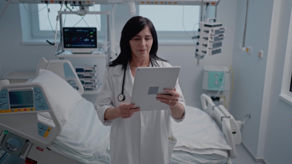 An older female doctor standing in hospital room, looking at tablet.