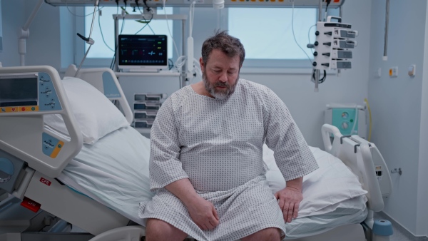 Male patient in a hospital gown sitting on hospital bed, waiting for surgery.