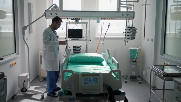 A doctor in a white coat checking a hospital room and filling out a medical form on a clipboard