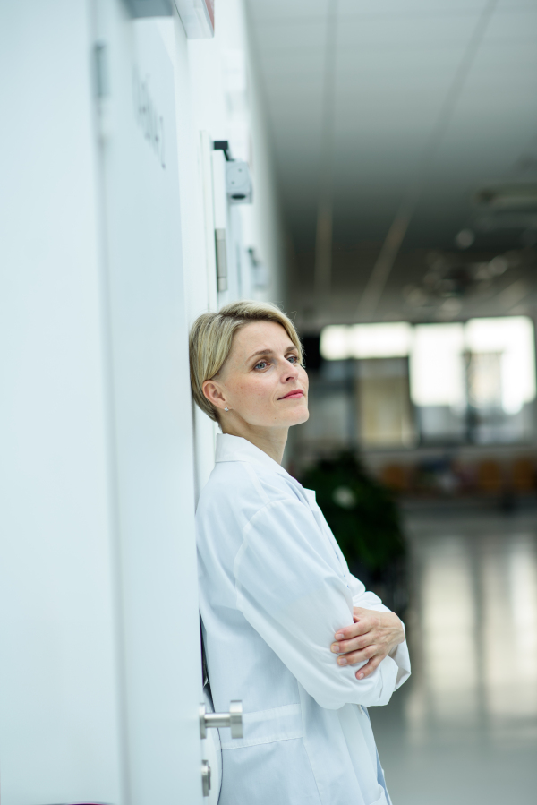 Tired female doctor leaning against wall in hospital hallway, side view.