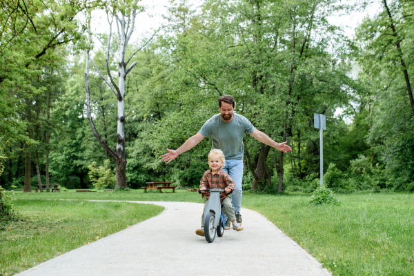 Toddler riding balance bike with dad cheering behind and running behind him.