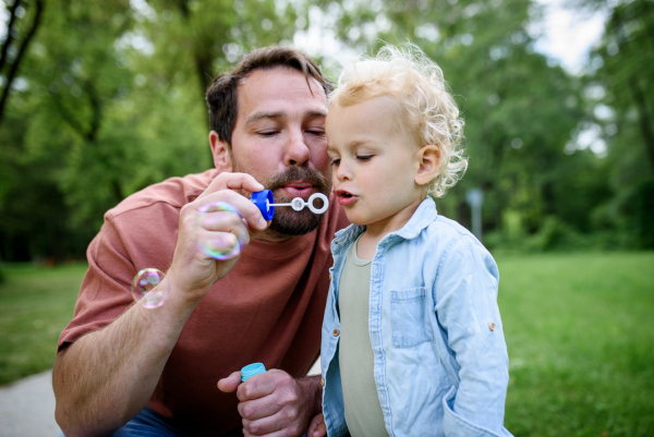 Father and son blowing soap bubbles in park. Toddler watching bubbles with amazement.
