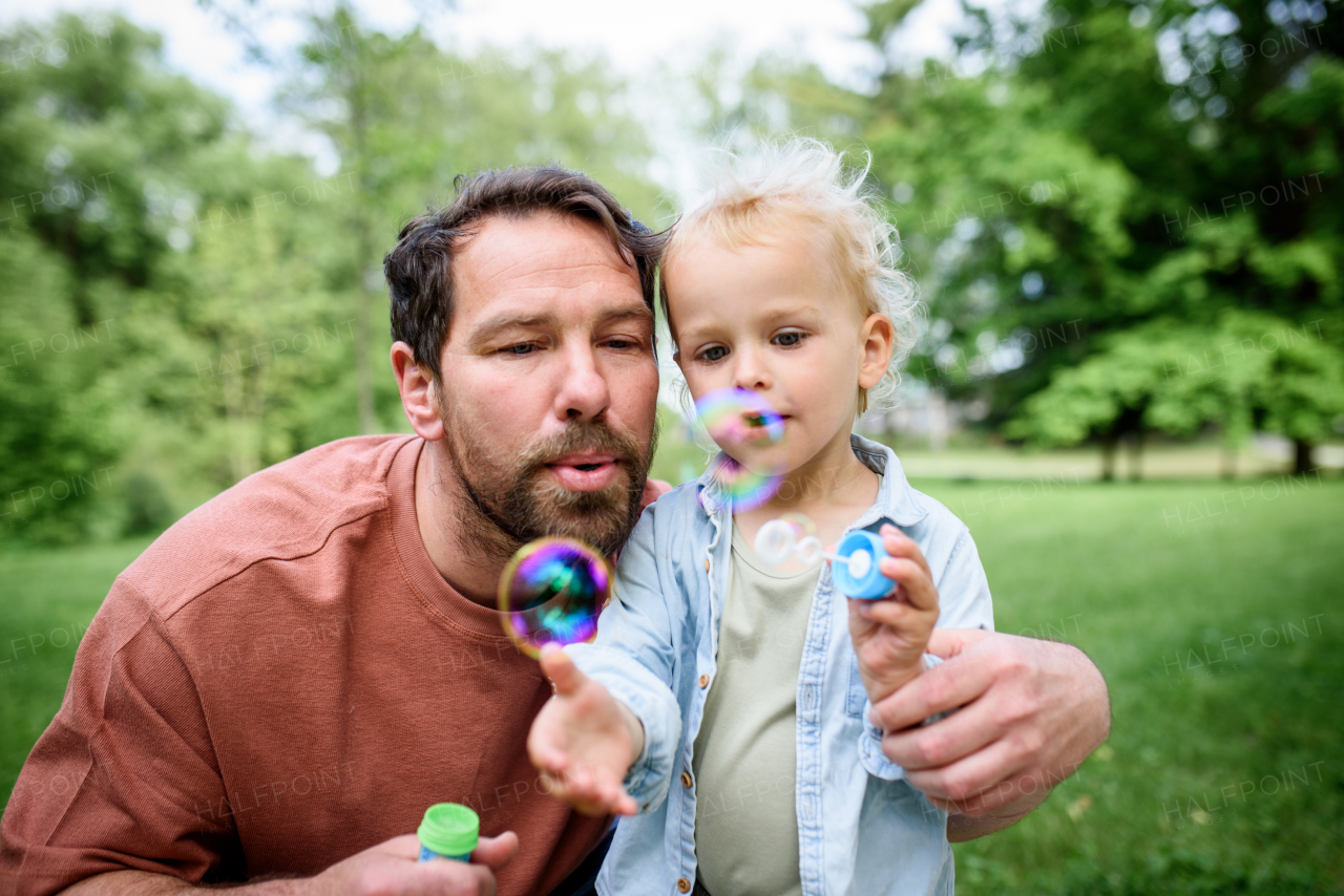 Father and son blowing soap bubbles in park. Toddler watching bubbles with amazement.