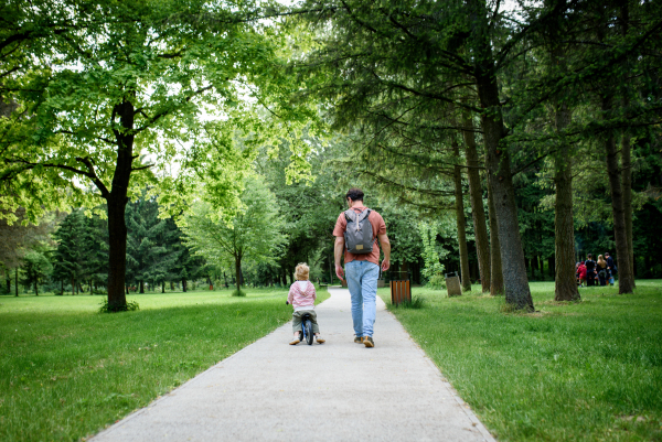 Rear view of father walking with his son balance bike across park