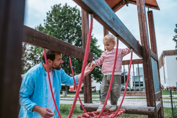 Toddler crossing playground obstacle with dad's support, weekend activity.