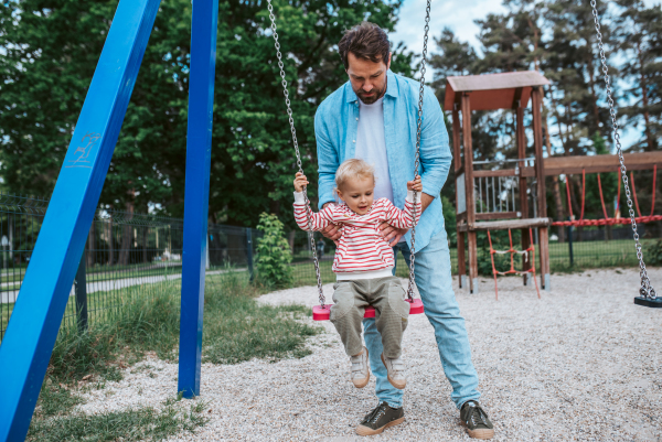 Father swinging small son on playground swing, weekend fun.