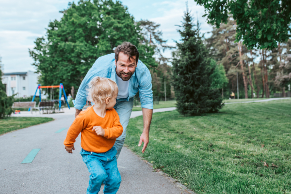 Father running with toddler son, chasing him for fun in park,