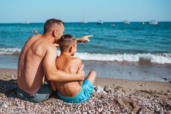 Rear view of son and father looking at sea, sitting on beach. Summer beach holiday.